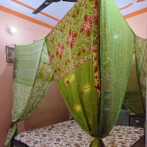 Könnte beinhalten: Ein Bett mit einer Matratze mit Blumenmuster und einem Baldachin aus grünem Stoff mit roten Blumendesigns. Der Baldachin ist über dem Bettrahmen drapiert und erzeugt einen gemütlichen und dekorativen Effekt. Der Bettrahmen ist hellfarbig mit einem dunkelgrauen Dekorationselement.