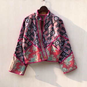 May include: A quilted jacket featuring a patchwork design with a mix of black, white, and red geometric patterns and floral motifs. The jacket has long sleeves, a mandarin collar, and is finished with bright pink piping along the edges.