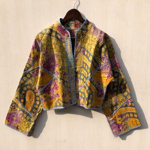 May include: A cropped jacket with a high collar, featuring a patchwork design in shades of yellow, purple, and gray. The jacket has a quilted texture and is trimmed with blue stitching. It is hanging on a wooden hanger.