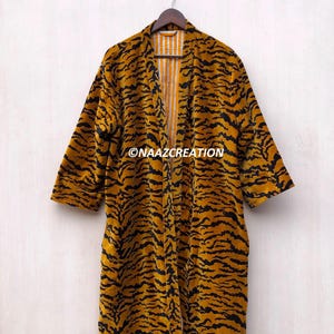 May include: A long-sleeved, open-front robe featuring a mustard yellow and black tiger stripe pattern. The garment has a straight silhouette and is displayed on a hanger. The text NAAZCREATION is printed in white on the front of the robe.