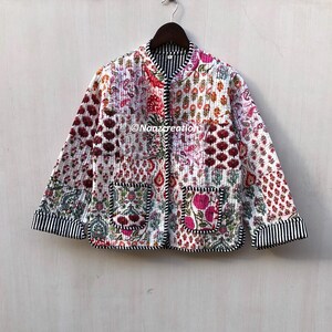 May include: A quilted jacket with a floral patchwork design in shades of red, pink, and white. The jacket has a mandarin collar, two front pockets, and black and white striped trim on the cuffs and hem. The jacket is hanging on a wooden hanger.
