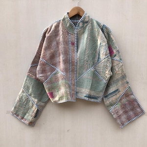 Könnte beinhalten: Eine kurz geschnittene Jacke aus gestepptem Stoff mit einem Patchwork-Muster in gedeckten Grün-, Braun- und Rosatönen. Das Kleidungsstück hat lange Ärmel, einen Stehkragen und sichtbare weiß-blaue Steppnähte entlang der Kanten und Nähte.