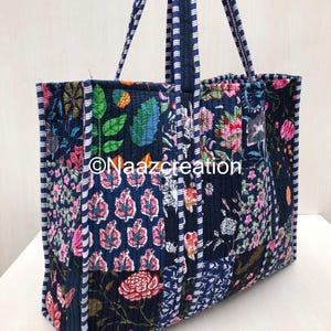 Puede incluir: Un bolso tote acolchado con fondo azul oscuro y un estampado floral y de hojas multicolor. El bolso tiene dos asas largas con un diseño de rayas azules y blancas. El texto Naazcreation está impreso en blanco en el centro.