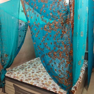 May include: A turquoise bed canopy with gold embroidery and embellishments. The canopy drapes over a bed with a floral patterned mattress. The bed frame is light-coloured with a decorative panel.
