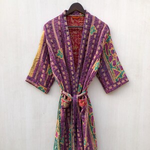May include: A purple, patterned robe with a matching belt, hanging on a wooden hanger. The robe features a mix of colors, including purple, yellow, green, and red. The robe is made of a soft, quilted fabric.