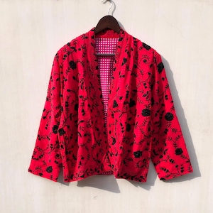 Puede incluir: Chaqueta roja con bordado floral negro, colgada de una percha de madera. El forro interior de la chaqueta es un estampado de cuadros rosas y blancos. La chaqueta tiene mangas largas y un corte holgado.