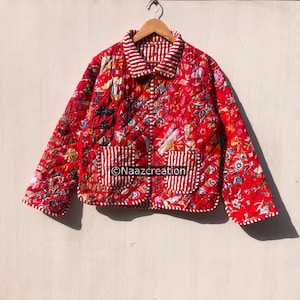 Puede incluir: Una chaqueta acolchada de tela roja vibrante con un patrón floral multicolor. La prenda incluye cuello, mangas largas y dos bolsillos delanteros, todos acentuados con un borde a rayas blancas y rojas. El texto Naazcreation es visible.