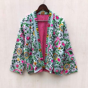 Könnte beinhalten: Weiß bestickte Jacke mit floralem Design in Rosa, Gelb und Grün. Die Jacke hat einen hellblauen Besatz und ein rosa-weiß kariertes Futter. Die Jacke hängt an einem Holzbügel.