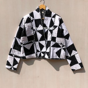 Könnte beinhalten: Schwarz-weiße Steppjacke mit geometrischem Muster. Die Jacke hat eine Mischung aus Dreiecken und Quadraten, mit Kragen und Taschen. Sie hängt an einem Holzkleiderbügel.