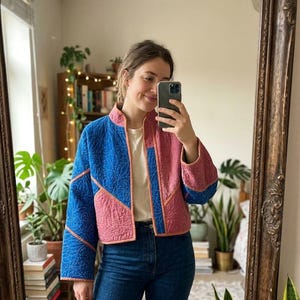 May include: A quilted jacket featuring a colour-blocked design in vibrant blue and dusty rose pink. The garment has a mandarin collar, long sleeves, and a textured, stitched pattern throughout. It is worn over a plain white t-shirt and blue jeans.