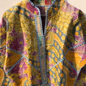 May include: A quilted jacket with a vibrant floral pattern in yellow, purple, and gray. The jacket features a mandarin collar and is trimmed with blue stitching. The design includes a patchwork style with various shapes and patterns.