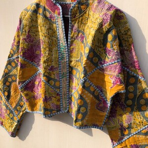 May include: A quilted jacket with a vibrant floral pattern in shades of yellow, purple, and blue. The jacket features a mandarin collar and is adorned with decorative stitching along the edges. The jacket is hanging on a wooden hanger.