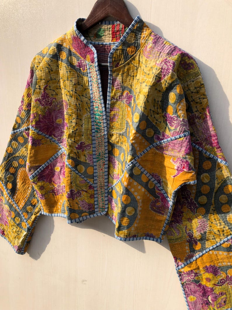 May include: A quilted jacket with a vibrant floral pattern in shades of yellow, purple, and blue. The jacket features a mandarin collar and is adorned with decorative stitching along the edges. The jacket is hanging on a wooden hanger.