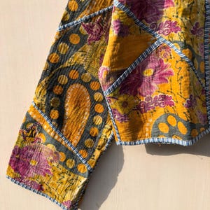 May include: Close-up of a patchwork jacket with a vibrant floral pattern in shades of yellow, purple, and gray. The jacket features a unique design with geometric shapes and blue trim, showcasing a bohemian style.
