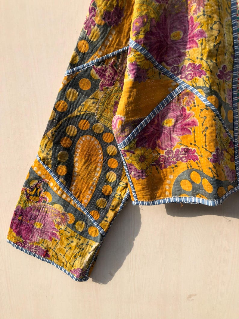 May include: Close-up of a patchwork jacket with a vibrant floral pattern in shades of yellow, purple, and gray. The jacket features a unique design with geometric shapes and blue trim, showcasing a bohemian style.