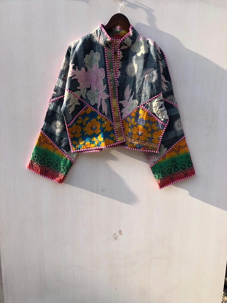 Puede incluir: Chaqueta patchwork con cuello alto y mangas largas. La chaqueta presenta una mezcla de estampados florales y geom&eacute;tricos en tonos azules, amarillos, rosas y verdes, con ribete rosa. Cuelga de una percha de madera.