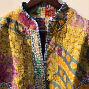 May include: A colorful, quilted jacket with a mandarin collar. The jacket features a vibrant mix of yellow, purple, and blue patterns. The edges are trimmed with a blue and white striped fabric. The jacket is hanging on a wooden hanger.
