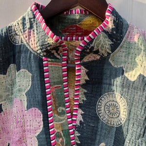 Puede incluir: Primer plano de una chaqueta acolchada con un estampado floral en tonos rosa, verde y blanco sobre un fondo azul oscuro. La chaqueta tiene cuello alto y est&aacute; ribeteada con tela a rayas rosas y blancas. La chaqueta est&aacute; colgada en una percha de madera.