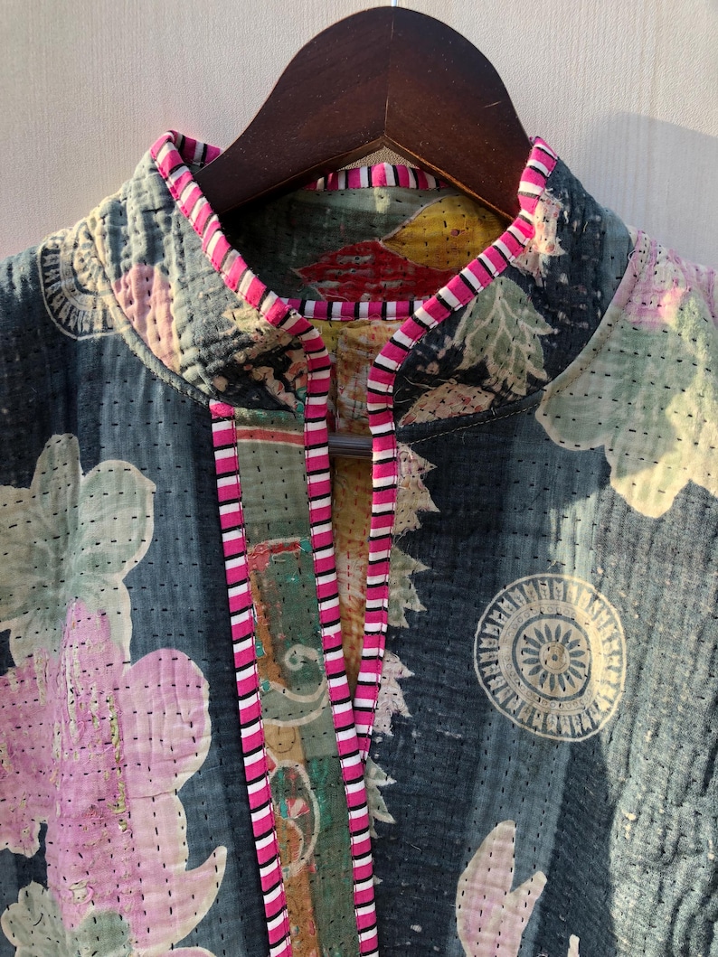 Puede incluir: Primer plano de una chaqueta acolchada con un estampado floral en tonos rosa, verde y blanco sobre un fondo azul oscuro. La chaqueta tiene cuello alto y est&aacute; ribeteada con tela a rayas rosas y blancas. La chaqueta est&aacute; colgada en una percha de madera.