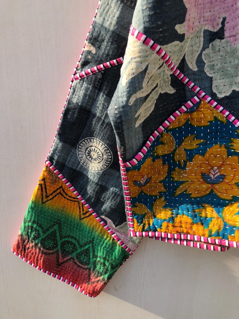 Puede incluir: Chaqueta patchwork con una variedad de patrones y colores. La chaqueta presenta una tela azul oscuro con un dise&ntilde;o circular blanco, un estampado floral en amarillo y azul, y una secci&oacute;n de colores del arco&iacute;ris. Ribetes rosas y blancos delinean las costuras.