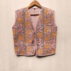 May include: A sleeveless quilted vest with a floral print in shades of pink, yellow, and gray. The vest has a tie closure at the neckline and a striped lining.