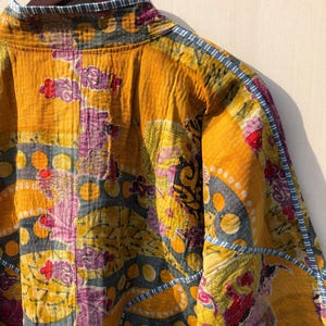 May include: A quilted jacket in shades of yellow, purple, and blue. The jacket features a patchwork design with floral and geometric patterns. The edges are trimmed with a blue and white striped fabric. The jacket is hanging on a wooden hanger.