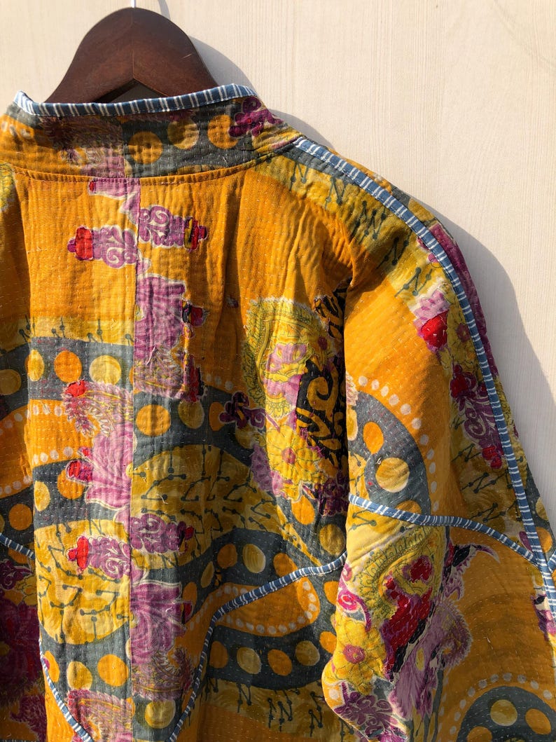 May include: A quilted jacket in shades of yellow, purple, and blue. The jacket features a patchwork design with floral and geometric patterns. The edges are trimmed with a blue and white striped fabric. The jacket is hanging on a wooden hanger.