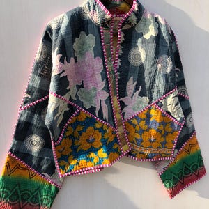 Puede incluir: Chaqueta patchwork con cuello alto, con estampados florales y geom&eacute;tricos en tonos azules, amarillos, rosas y verdes. La chaqueta tiene una textura acolchada y est&aacute; ribeteada con costuras rosas. Cuelga de una percha de madera.