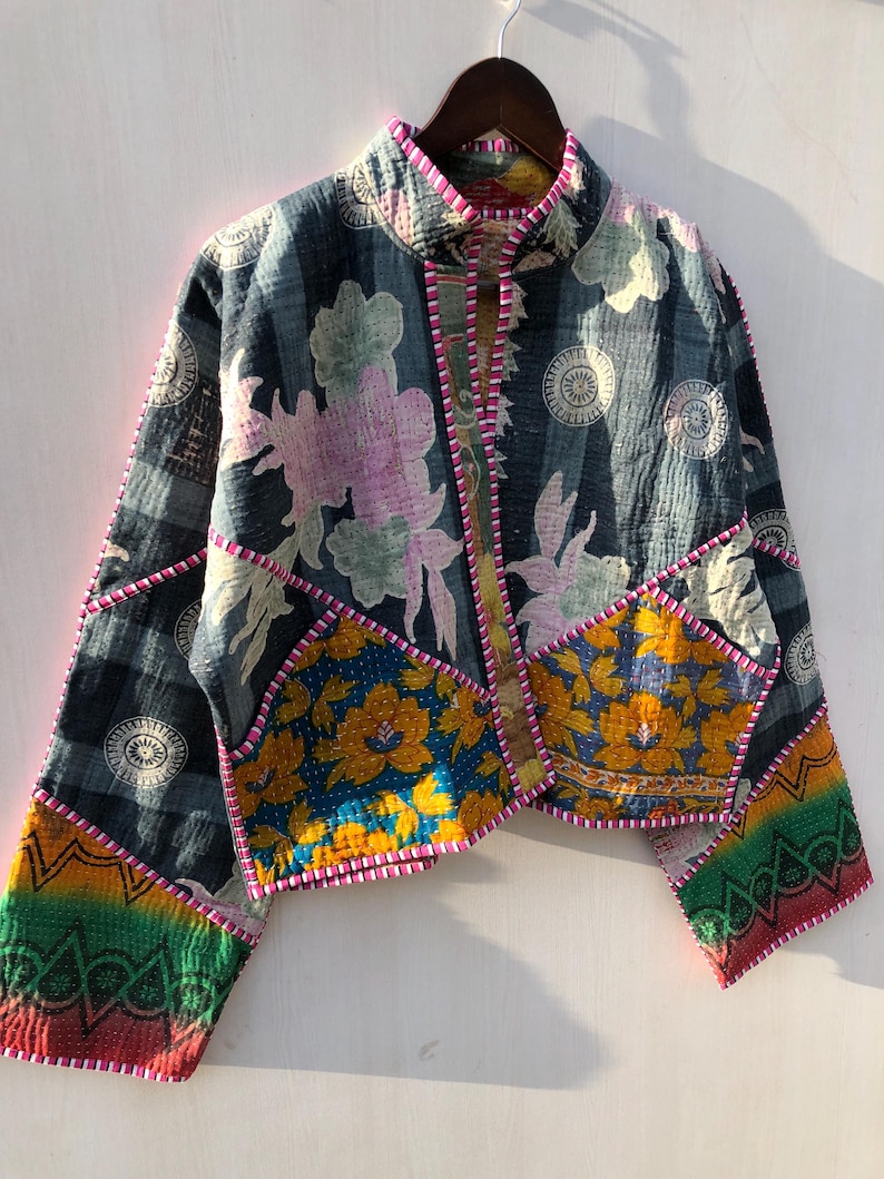 Puede incluir: Chaqueta patchwork con cuello alto, con estampados florales y geom&eacute;tricos en tonos azules, amarillos, rosas y verdes. La chaqueta tiene una textura acolchada y est&aacute; ribeteada con costuras rosas. Cuelga de una percha de madera.