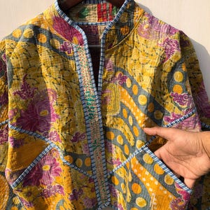 May include: A quilted jacket with a vibrant patchwork design. The jacket features a mix of yellow, purple, and gray floral and geometric patterns. It has a mandarin collar, pockets, and is trimmed with blue and white striped fabric.