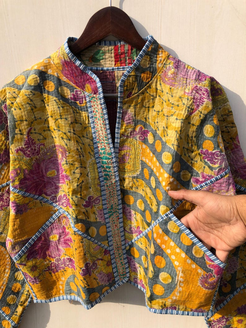May include: A quilted jacket with a vibrant patchwork design. The jacket features a mix of yellow, purple, and gray floral and geometric patterns. It has a mandarin collar, pockets, and is trimmed with blue and white striped fabric.