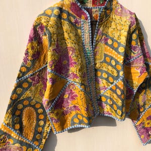 May include: A cropped jacket with a vibrant patchwork design. The jacket features a high collar and long sleeves. The fabric is a mix of yellow, purple, and blue floral and geometric patterns. The edges are trimmed with blue stitching. The jacket is hanging on a wooden hanger.