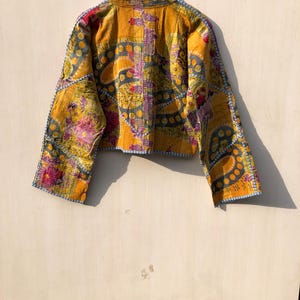 May include: A cropped, long-sleeve jacket with a vibrant, patchwork design. The jacket features a yellow base with floral and geometric patterns in purple, gray, and red. The edges are trimmed with blue stitching, and the jacket is hanging on a wooden hanger.