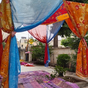 Puede incluir: Un dosel colorido hecho de tela drapeada en tonos naranja, rojo, azul y amarillo. La tela tiene diseños florales y geométricos. El dosel está instalado al aire libre, con vegetación en el fondo.