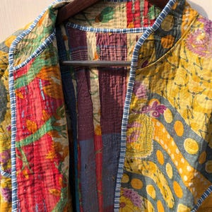 May include: A colorful patchwork jacket with a variety of patterns and colors, including red, yellow, and blue. The jacket is hanging on a wooden hanger. The edges are trimmed with a blue and white striped fabric.