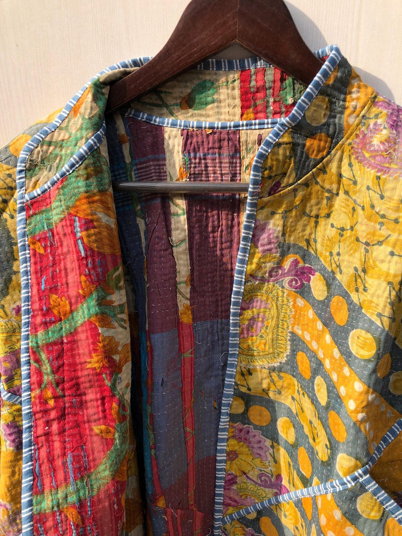 May include: A colorful patchwork jacket with a variety of patterns and colors, including red, yellow, and blue. The jacket is hanging on a wooden hanger. The edges are trimmed with a blue and white striped fabric.