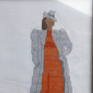 May include: Cross-stitch embroidery of a woman wearing a gray coat and an orange dress with a hat.