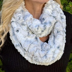 May include: A soft, faux fur infinity scarf in shades of white, gray, and black. The plush scarf is wrapped around a person's neck, with the black sweater providing a contrasting background. The scarf appears to be made of a fluffy, textured material.