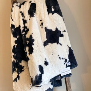 May include: A black and white cow print plush blanket with a Minky Luxe Co. tag attached. The blanket is folded and draped over a wooden ladder.