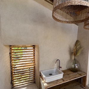 May include: A bathroom interior with a white rectangular sink and copper tap on a wooden shelf. A glass vase with dried plants is to the right. Two woven pendant lights hang from the ceiling. The window has wooden slats.