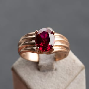 May include: A gold ring with a large, oval-shaped, red gemstone set in the center. The ring has a textured band with multiple grooves.