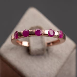 Può includere: Anello in oro rosa con cinque pietre preziose rosse ovali. Le pietre sono disposte in fila sulla parte superiore dell'anello. L'anello è presentato su uno sfondo neutro, che mette in risalto le gemme.