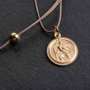 Könnte beinhalten: Goldfarbene Anhänger Halskette mit einem runden Medaillon mit einem detaillierten Relief einer religiösen Figur. Der Anhänger hängt an einer dünnen, beigefarbenen Kordel mit einer goldenen Perle. Die Halskette wird vor dunklem Hintergrund präsentiert.