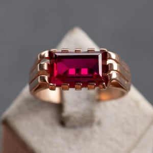 Op de afbeelding: Een goudkleurige ring met een grote, rechthoekige, dieprode edelsteen. De ring heeft een klassiek ontwerp met een band die naar de bovenkant breder wordt, en de edelsteen is gezet met kleine, vierkante accenten. De ring wordt op een neutrale ondergrond gepresenteerd.