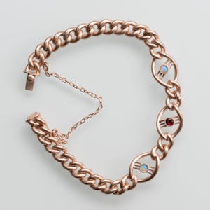 Może przedstawiać: Bransoletka z łańcuszkiem z różowego złota z trzema ozdobnymi owalnymi ogniwami. Każde owalne ogniwo ma mały kamień szlachetny, czerwony lub niebiesko-biały. Bransoletka jest rozpięta, pokazując łańcuszek i łańcuszek zabezpieczający.