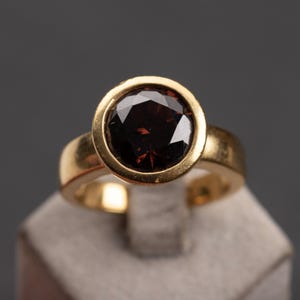 May include: A gold ring featuring a large, faceted, dark red gemstone. The gemstone is set in a bezel setting, and the band is a simple, polished gold. The ring is displayed on a gray surface against a dark gray background.