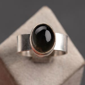 Könnte beinhalten: Ein silberner Ring mit einem schwarzen ovalen Edelstein, der in einer Zargenfassung gefasst ist.