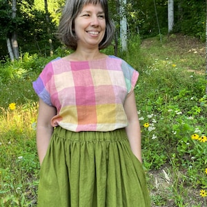 Pode incluir: Uma blusa xadrez colorida em tons de rosa, amarelo e azul combinada com uma saia longa verde-oliva. O conjunto é feito de um tecido macio e fluido, perfeito para um ambiente casual ao ar livre. A blusa tem decote quadrado e mangas curtas.