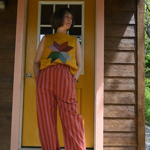 May include: A woman wearing a yellow sleeveless top with a colorful star design and red and pink striped pants. The outfit is casual and the woman is standing on a wooden porch in front of a yellow door.