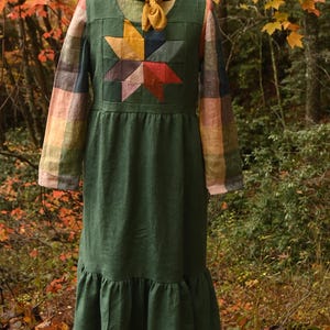 May include: A green dress with a starburst design on the bodice. The dress has long sleeves with a plaid pattern and a ruffled hem. A yellow scarf is tied around the neckline. The dress is displayed on a mannequin in a forest setting.